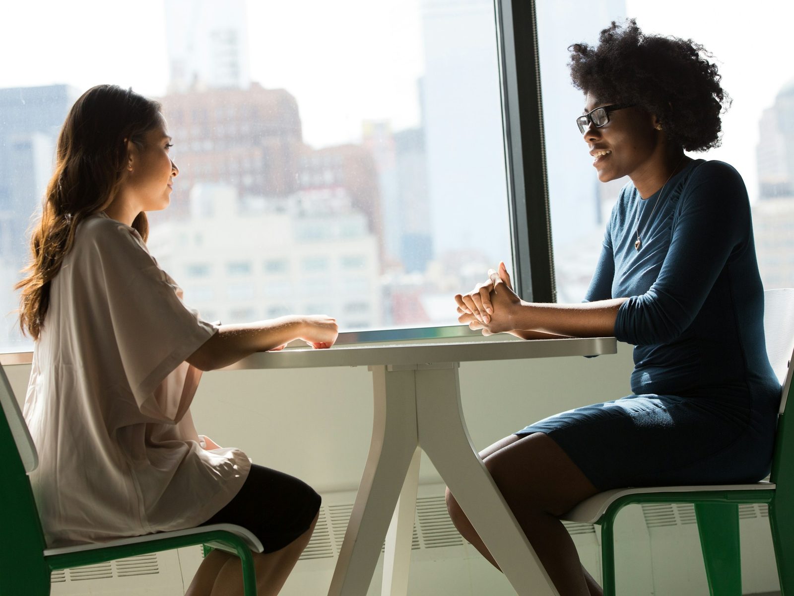 Two colleagues in conversation across a table by a bright office window.