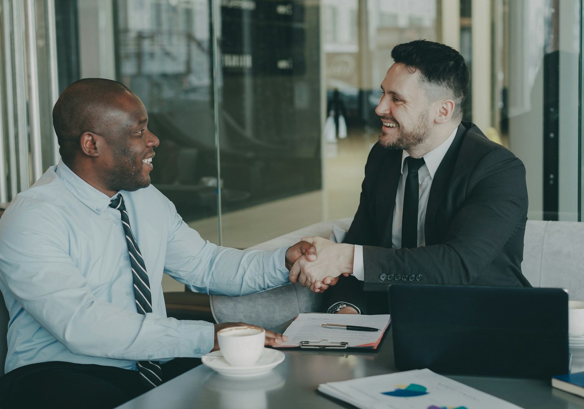 Two professionals shaking hands across a table in a bright modern office.